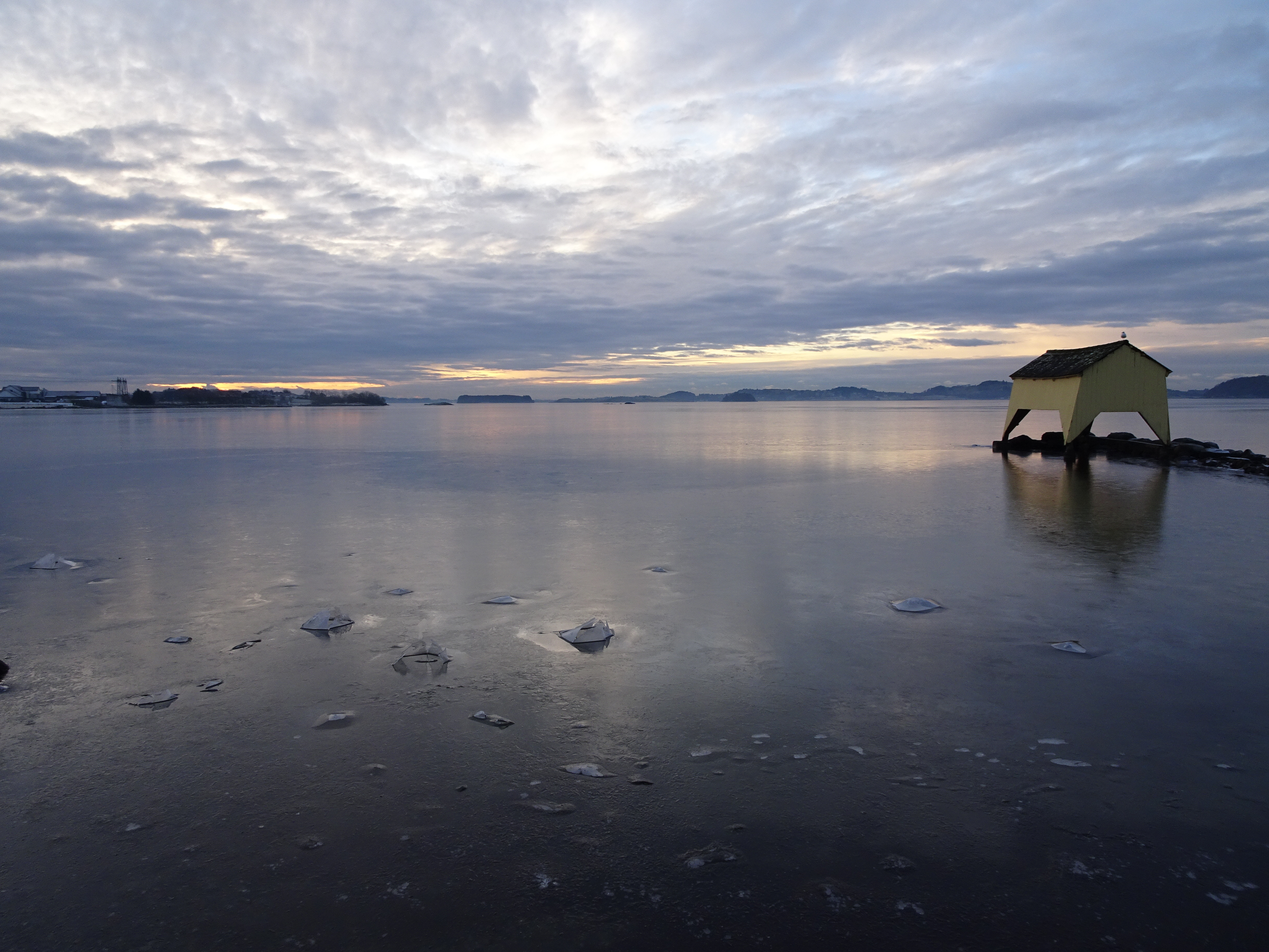 A view across Hafrsfjord in the winter with a yellow boathouse in the middle ground and the water frozen in the foreground.