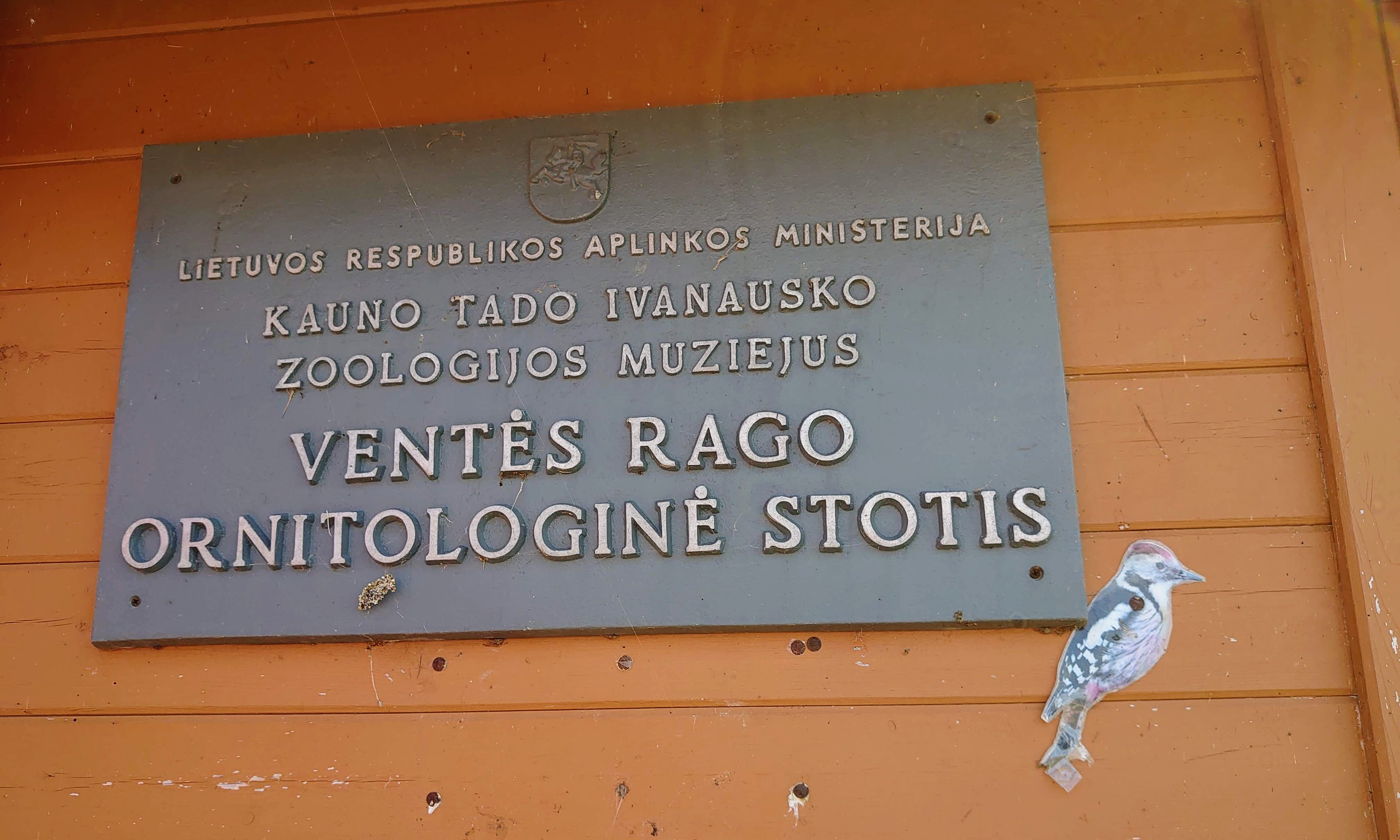 The sign for the Ventė Cape Ornithological Station on a wooden wall with a woodpecker sticker to the bottom right of it.
