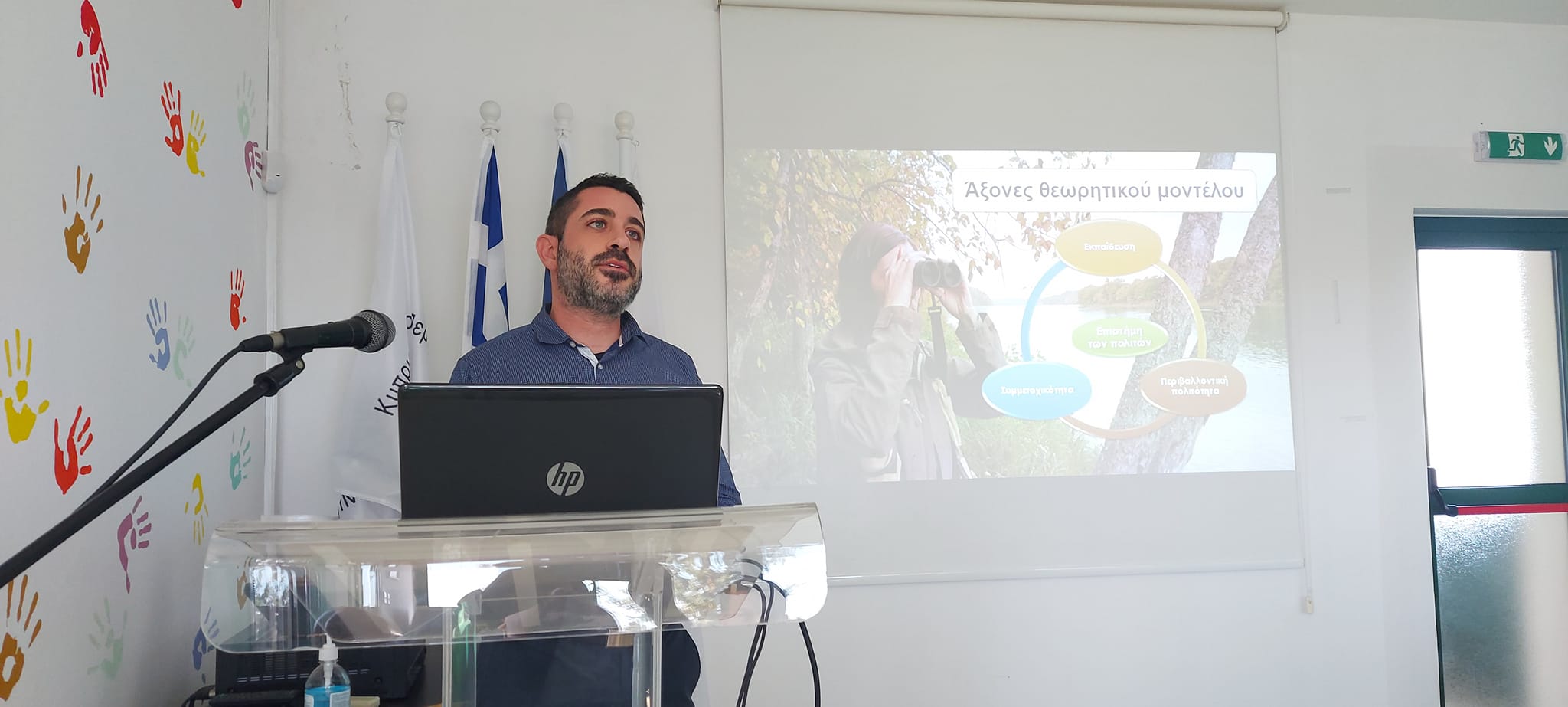 A lecturer stands at a lectern. behind him is a Powerpoint slide with a circular diagram on it. The text is in Greek